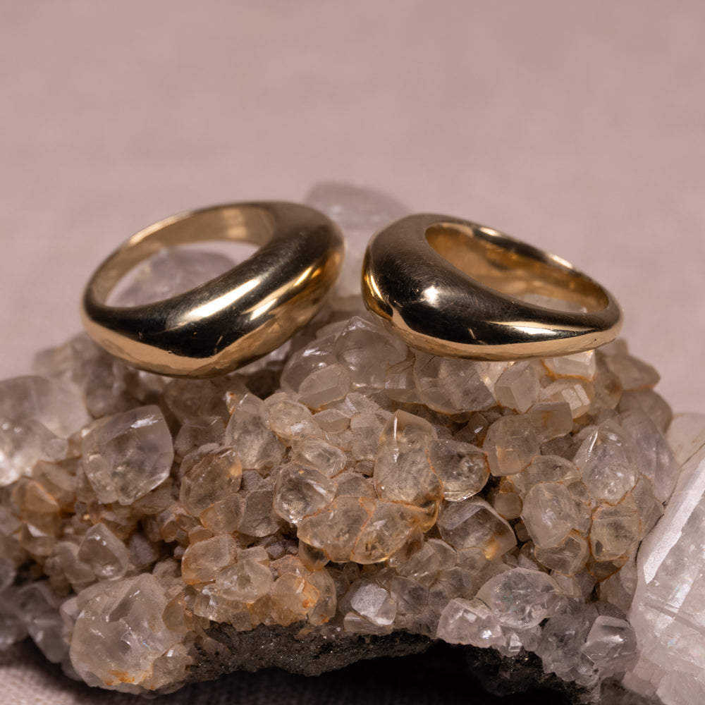 Two polished gold rings with a smooth, rounded organic shape resting on a cluster of clear quartz crystals against a neutral beige background.