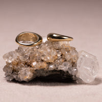 Two polished gold rings, one a simple band and the other a teardrop-shaped ring, displayed on a cluster of clear quartz crystals against a neutral beige background.