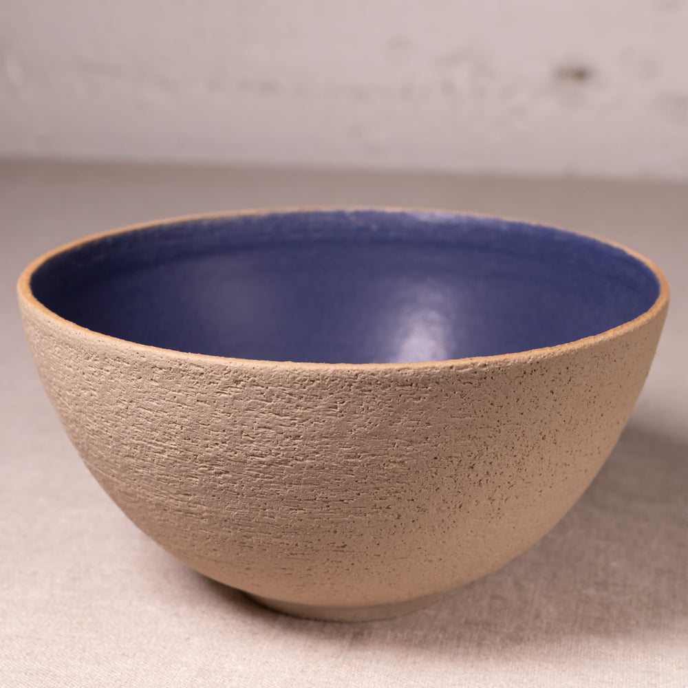 A handmade ceramic bowl with a textured beige exterior and a smooth, glossy blue interior, set against a neutral light gray background.
