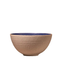 A handmade ceramic bowl with a textured beige exterior and a glossy dark blue interior, set against a rustic, light-colored brick wall background.