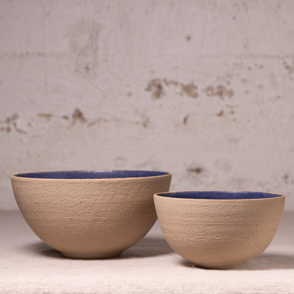 Two handmade ceramic bowls with a textured beige exterior and a deep blue glazed interior, displayed against a rustic, distressed light gray wall.
