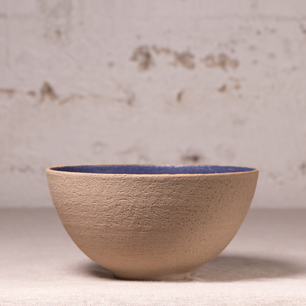 A handmade ceramic bowl with a textured beige exterior and a contrasting dark blue glazed interior, set against a rustic, light-colored brick wall background.