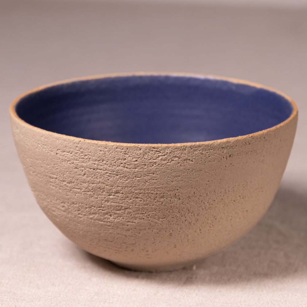 A handmade ceramic bowl with a textured beige exterior and a smooth, glossy dark blue interior, photographed on a neutral beige background.