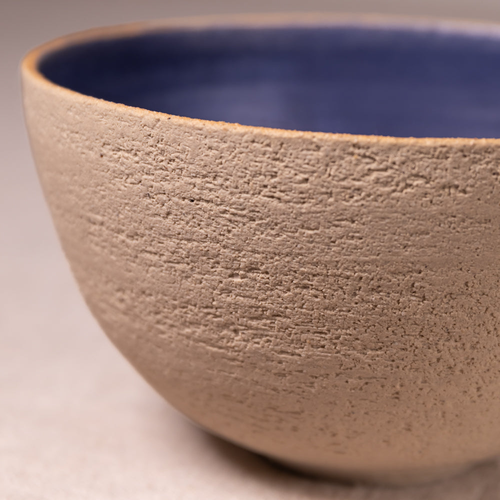 Close-up of a handmade ceramic bowl with a textured beige exterior and a smooth dark blue interior.
