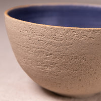 Close-up of a handmade ceramic bowl with a textured beige exterior and a smooth dark blue interior.