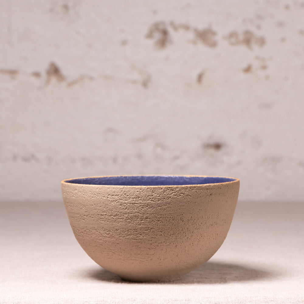 A handmade ceramic bowl with a textured beige exterior and a contrasting blue glazed interior, set against a rustic, light-colored plaster wall.