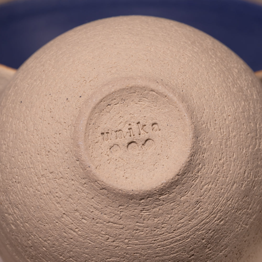 Close-up of the bottom of a textured ceramic bowl or vase, showing a stamped maker's mark reading 'umika' in a circular recess, with a dark blue background visible around the edges.