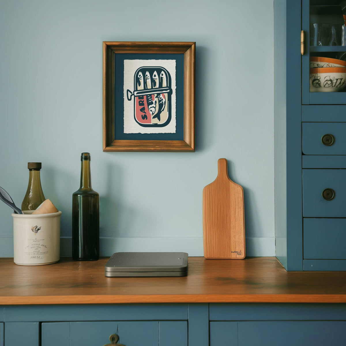 Kitchen counter with a wooden cutting board, bottles, and a framed artwork on a light blue wall.
