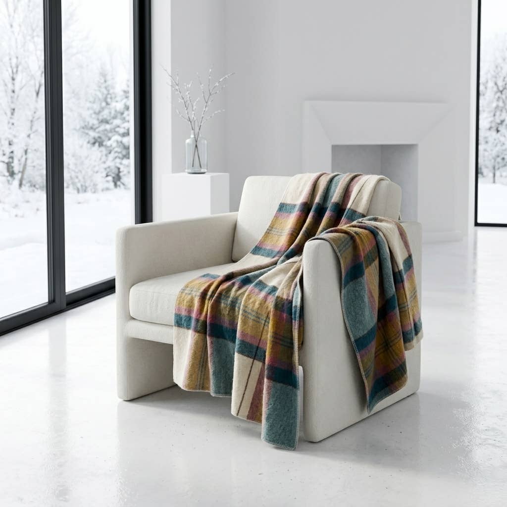 A multicolor plaid throw blanket in shades of blue, green, yellow, and beige is draped over a modern off-white armchair in a bright, minimalist room with large windows showing a snowy outdoor view.