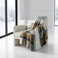 A multicolor plaid throw blanket in shades of blue, green, yellow, and beige is draped over a modern off-white armchair in a bright, minimalist room with large windows showing a snowy outdoor view.