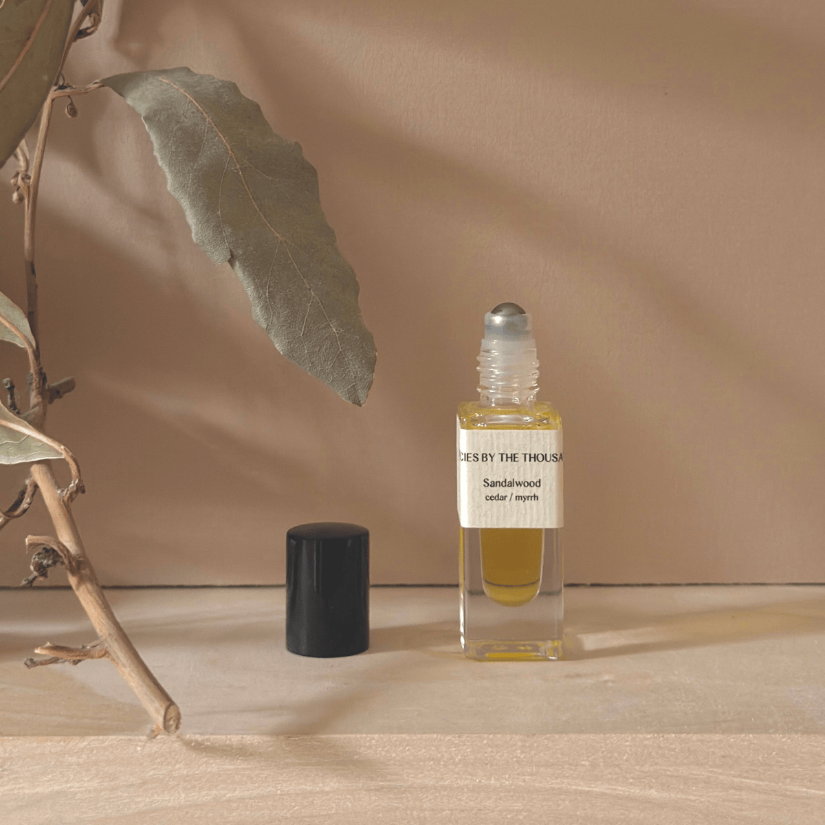 Small glass rollerball bottle with sandalwood essential oil on a wooden surface with a leaf and branch.