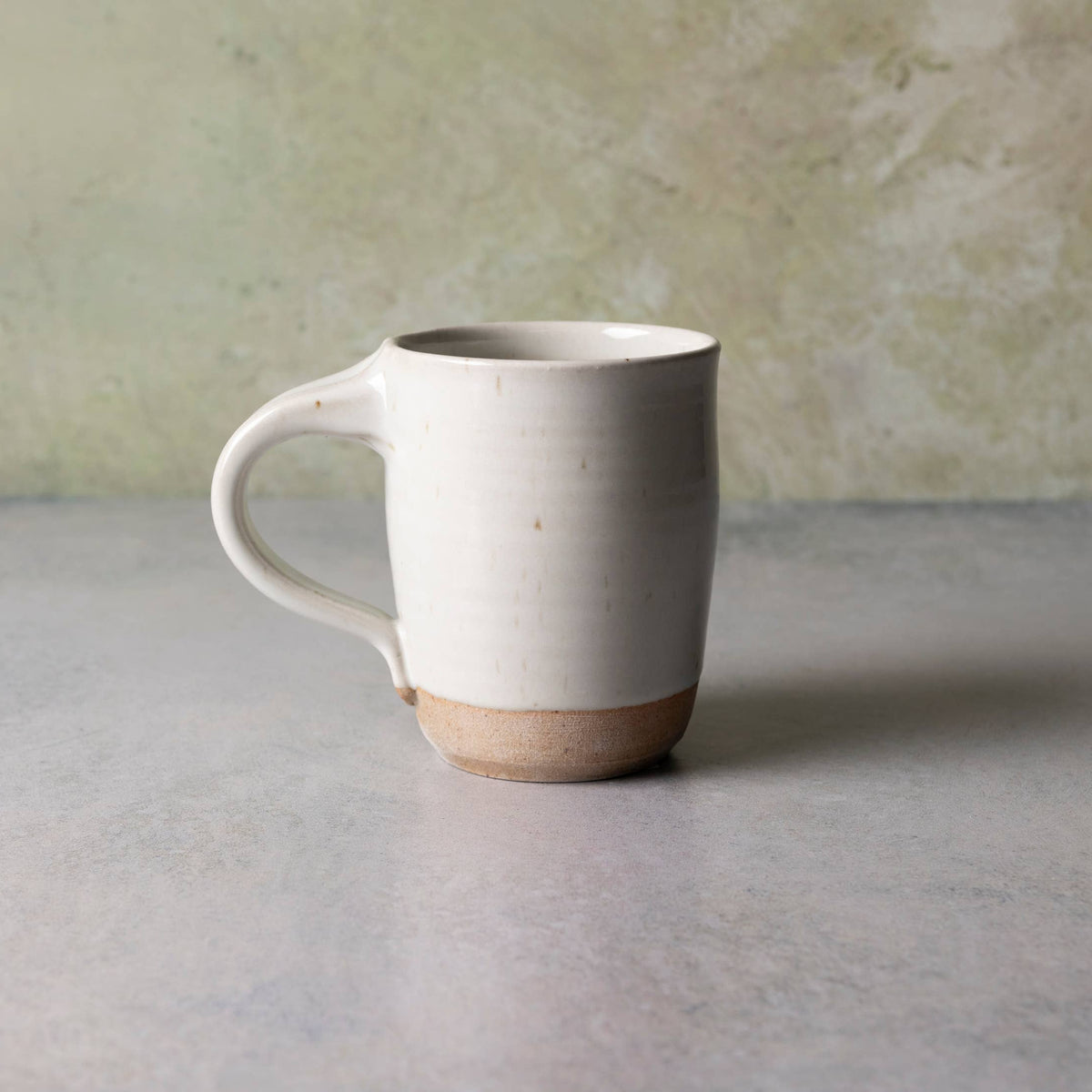 A handmade ceramic mug with a matte off-white body, a natural tan unglazed base, and a curved handle, set against a textured light gray and green background.
