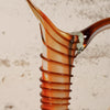 Close-up of a hand-blown glass vase with a flared rim and a spiral ribbed stem, featuring amber, orange, and clear glass tones against a textured beige background.