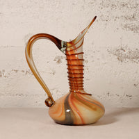 A decorative hand-blown glass pitcher with a curved handle, ribbed neck, and tapered spout, featuring marbled amber, orange, cream, and dark brown tones, set against a textured off-white wall.