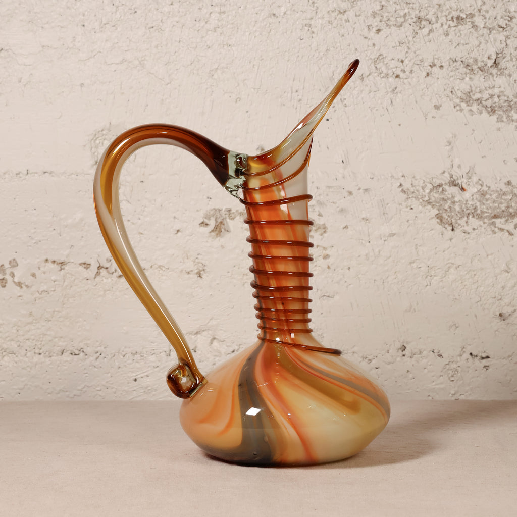 A decorative hand-blown glass pitcher with a curved handle, ribbed neck, and tapered spout, featuring marbled amber, orange, cream, and dark brown tones, set against a textured off-white wall.