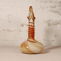 A hand-blown glass vase with a bulbous base, a ribbed neck, and a narrow pointed top, featuring marbled swirls of cream, orange, and brown, set against a textured off-white wall.