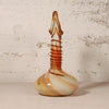 A hand-blown glass vase with a bulbous base, a ribbed neck, and a narrow pointed top, featuring marbled swirls of cream, orange, and brown, set against a textured off-white wall.