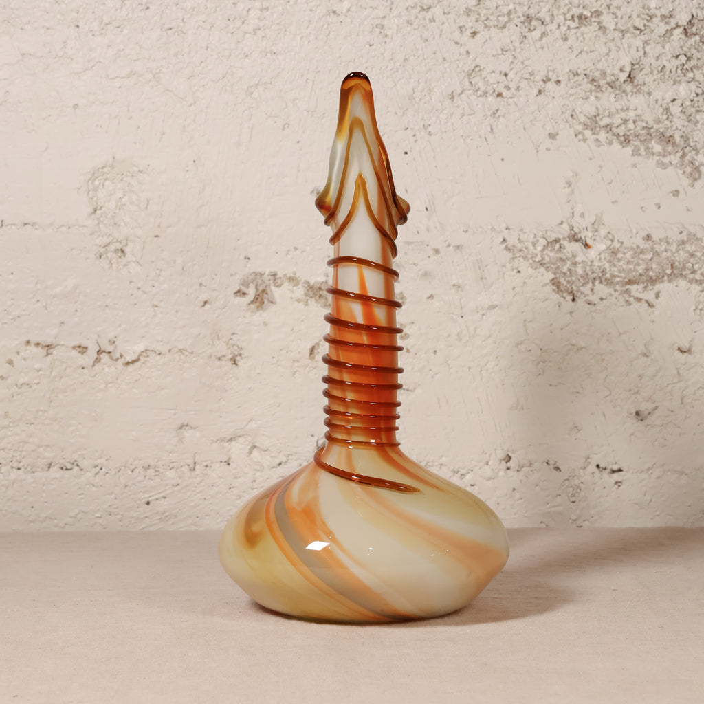 A hand-blown glass vase with a bulbous base, a ribbed neck, and a narrow pointed top, featuring marbled swirls of cream, orange, and brown, set against a textured off-white wall.