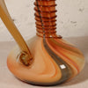 A close-up of a hand-blown glass vessel with a bulbous base, a coiled neck, and a curved stem, featuring marbled swirls of amber, orange, peach, and translucent cream against a light textured wall.