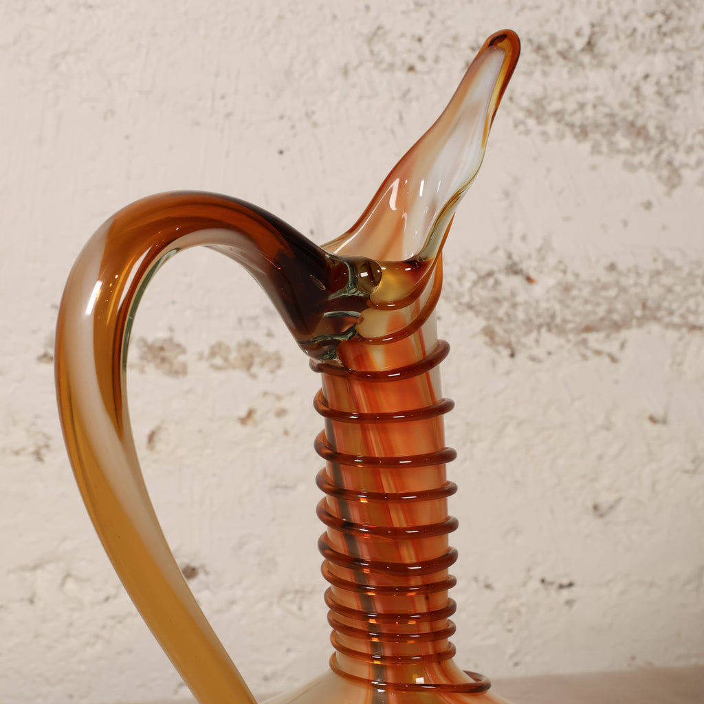 A close-up of a decorative amber glass pitcher with a curved handle and ribbed neck, set against a textured off-white wall.