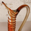 A close-up of a hand-blown glass decanter with a curved handle and a ribbed neck, featuring amber, orange, and cream glass swirls against a textured light beige background.