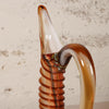 A close-up of a hand-blown glass decanter with a ribbed stem and curved handle, featuring amber, brown, and cream glass tones, set against a textured off-white wall.