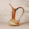 A decorative blown-glass pitcher with a ribbed neck, curved handle, and bulbous base, featuring amber, orange, cream, and translucent tones against a textured off-white wall.