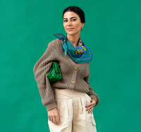 Woman wearing a brown sweater, blue scarf, and holding a green bag against a teal background