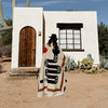 Person wearing a patterned blanket in front of a white building with a wooden door in a desert setting.