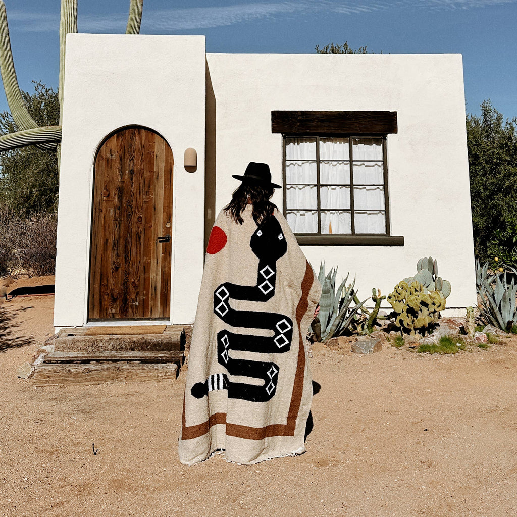 Person wearing a patterned blanket in front of a white building with a wooden door in a desert setting.