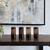 Four cylindrical glass containers on a wooden table with a decorative plant and abstract painting in the background.