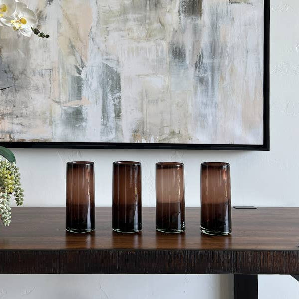 Four cylindrical glass containers on a wooden table with a decorative plant and abstract painting in the background.