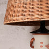 Close-up of a woven natural rattan lampshade mounted on a black lamp base over a white ceramic lamp with a brown handle and red floral decoration, set against a textured off-white wall.