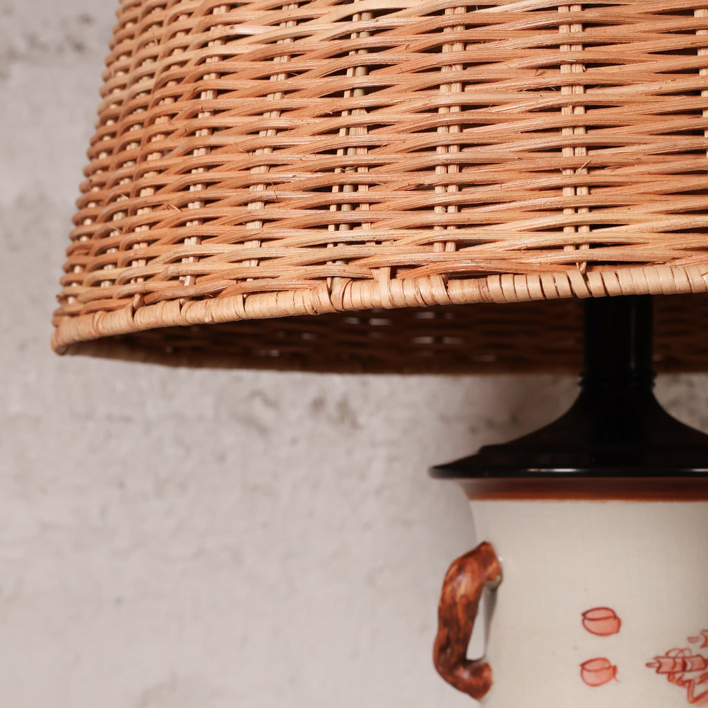 Close-up of a woven natural rattan lampshade mounted on a black lamp base over a white ceramic lamp with a brown handle and red floral decoration, set against a textured off-white wall.