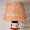 A table lamp with a large natural woven rattan shade and a ceramic base featuring a white body, reddish-brown accents, and small red floral details, set against a textured light gray wall.