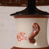 Close-up of a ceramic table lamp base with a cream-colored body, a glossy dark brown neck and lid, a sculpted reddish-brown handle, and a red hand-painted design on the side.