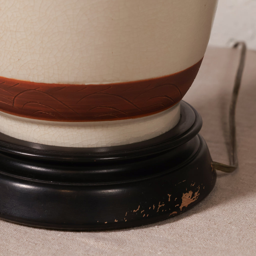 Close-up of a ceramic lamp base featuring a cream-colored body with a brown band, set on a black round wooden stand.