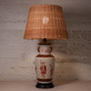 A table lamp with a natural wicker shade and a ceramic vase base decorated with red figures and motifs, set against a textured concrete wall.