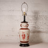 A vintage-style table lamp with a cream-colored ceramic vase body decorated with red figures and motifs, a black neck and base, and a brass-colored light fixture with a clear bulb.