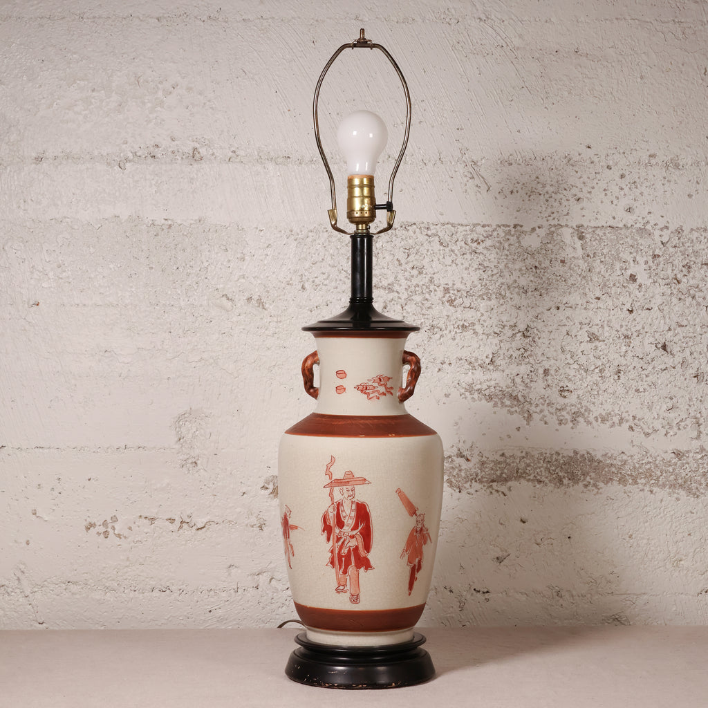 A vintage-style table lamp with a cream-colored ceramic vase body decorated with red figures and motifs, a black neck and base, and a brass-colored light fixture with a clear bulb.