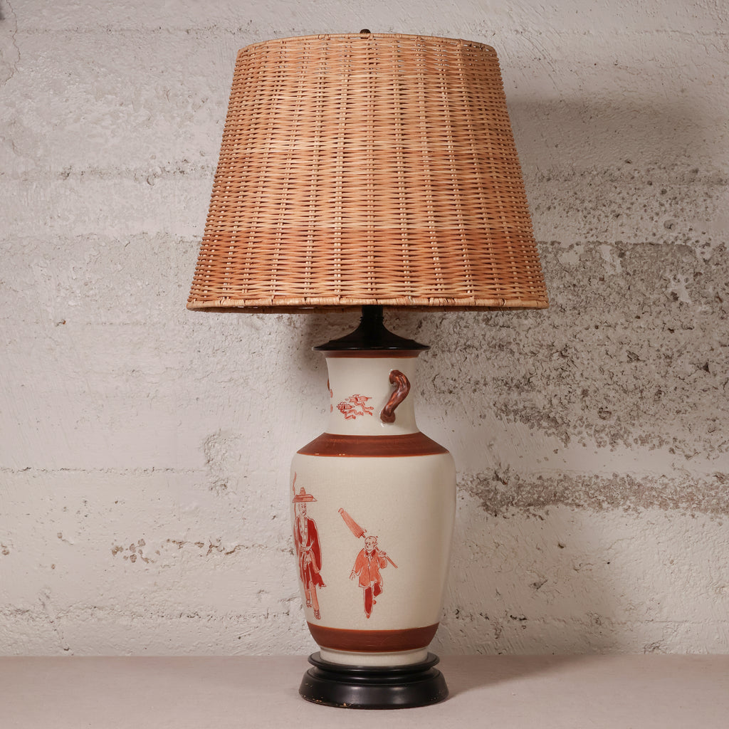 A vintage-style table lamp with a natural woven wicker shade and a ceramic base decorated with red figures on a cream background, set against a textured light gray wall.