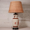 A vintage-style table lamp with a natural woven rattan shade, a cream-colored ceramic base decorated with reddish-brown patterns, and a dark metal foot ring, set against a textured light gray wall.