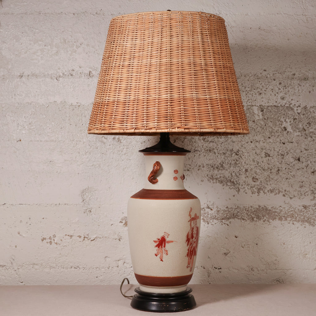 A vintage-style table lamp with a natural woven rattan shade, a cream-colored ceramic base decorated with reddish-brown patterns, and a dark metal foot ring, set against a textured light gray wall.