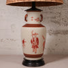 A ceramic table lamp with a cream-colored body decorated with red figures and motifs, featuring a woven wicker shade and a dark base.