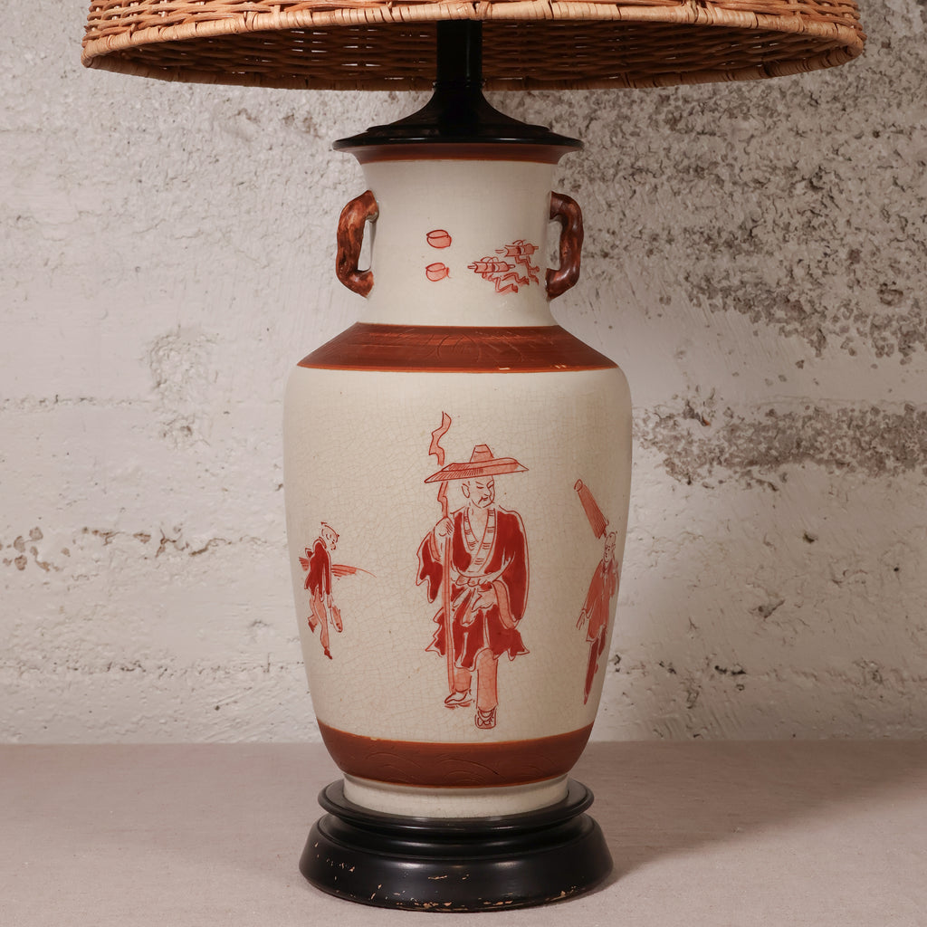 A ceramic table lamp with a cream-colored body decorated with red figures and motifs, featuring a woven wicker shade and a dark base.