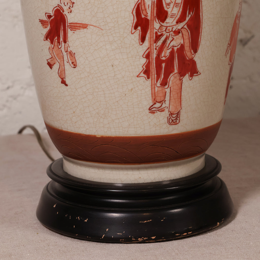 A close-up of a ceramic table lamp base featuring a cream-colored glaze with red hand-painted figures and a brown band, mounted on a round black wooden stand.