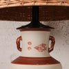 A close-up of a ceramic table lamp base with a cream-colored body, reddish-brown handles and bands, and a black neck supporting a woven wicker shade.