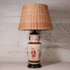 A vintage table lamp featuring a natural woven wicker shade and a ceramic base with red hand-painted figures on a cream background, set against a textured light gray wall.
