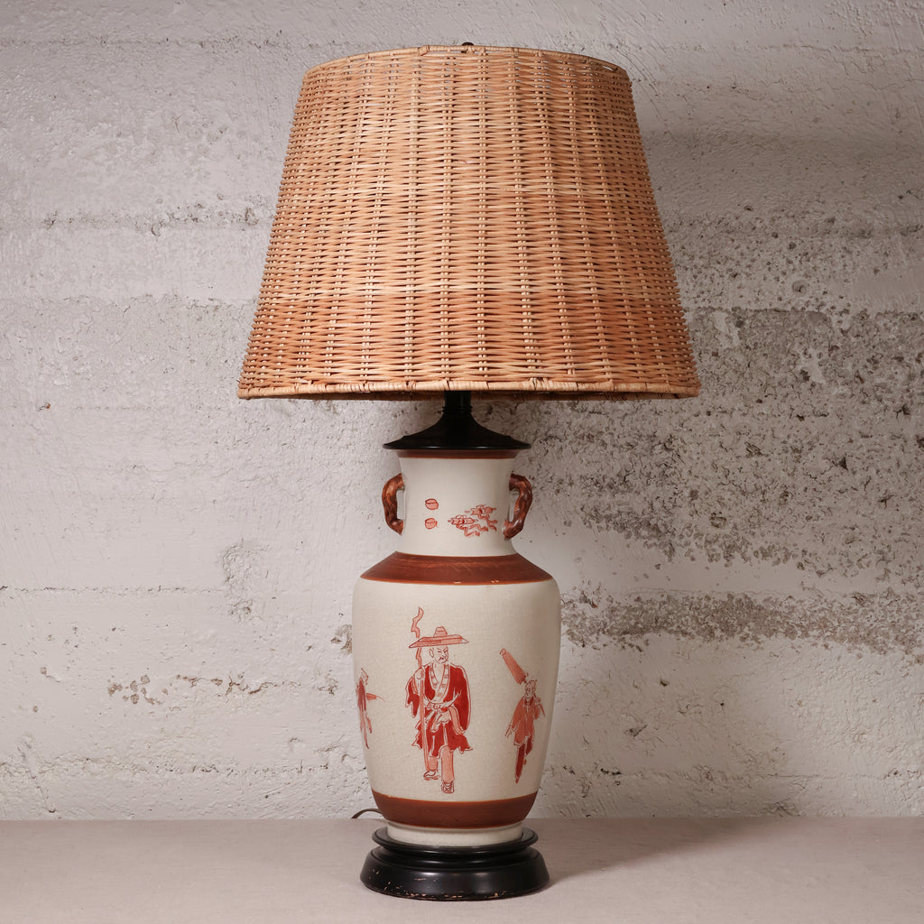 A vintage table lamp featuring a natural woven wicker shade and a ceramic base with red hand-painted figures on a cream background, set against a textured light gray wall.