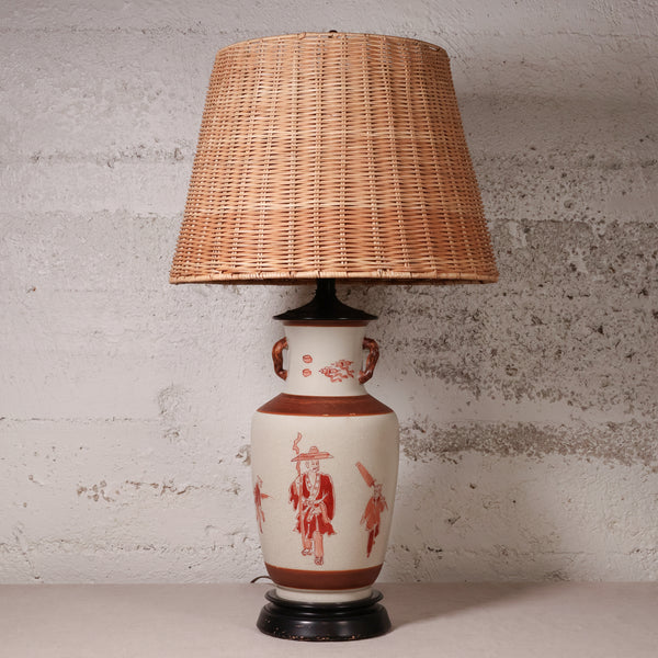 A vintage table lamp featuring a natural woven wicker shade and a ceramic base with red hand-painted figures on a cream background, set against a textured light gray wall.